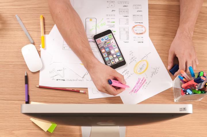 This is an image of a persons hands' highlighting somthing on a piece of paper. The paper is on a table with other stationary and more papers. There is also a desktop screen on the table